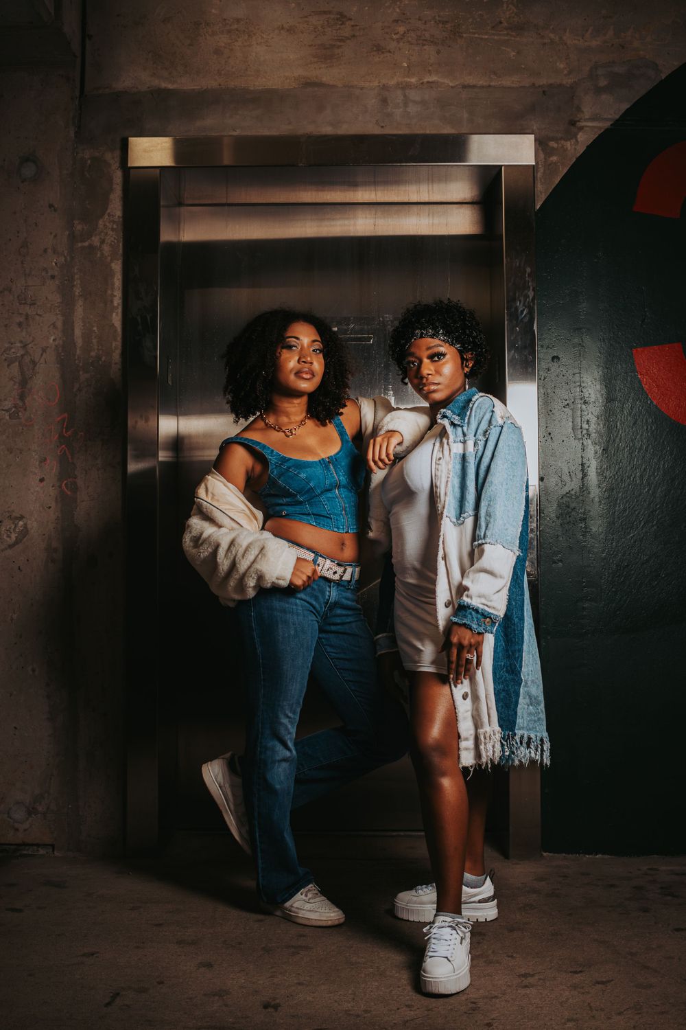 Two models pose casually in denim and blue outfits against an industrial elevator setting.