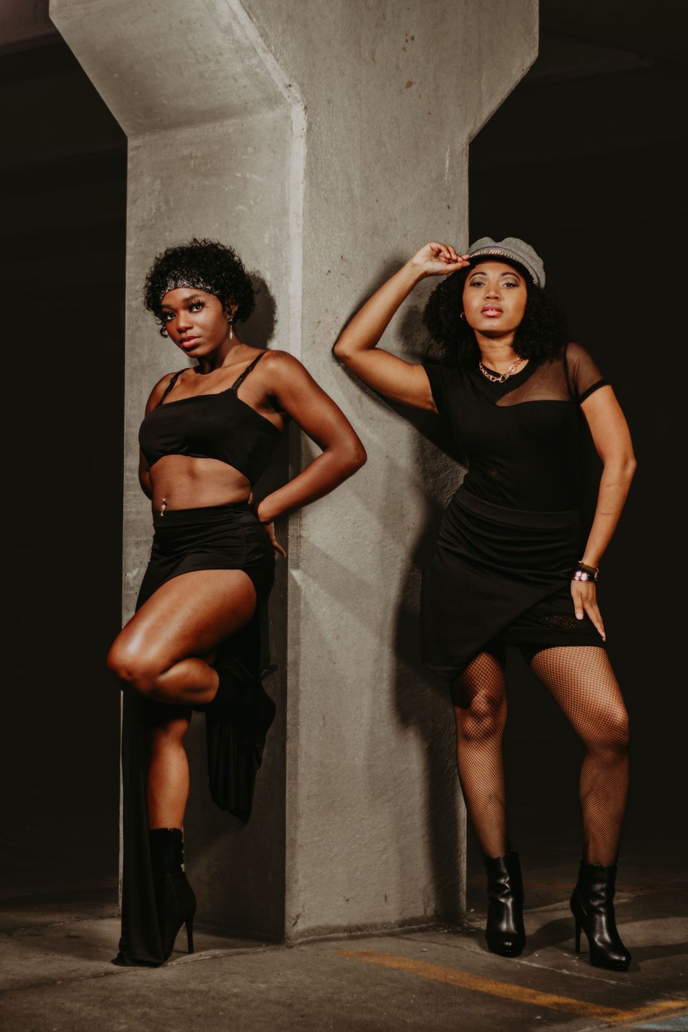Two models in black outfits strike confident poses against concrete pillar backdrop.