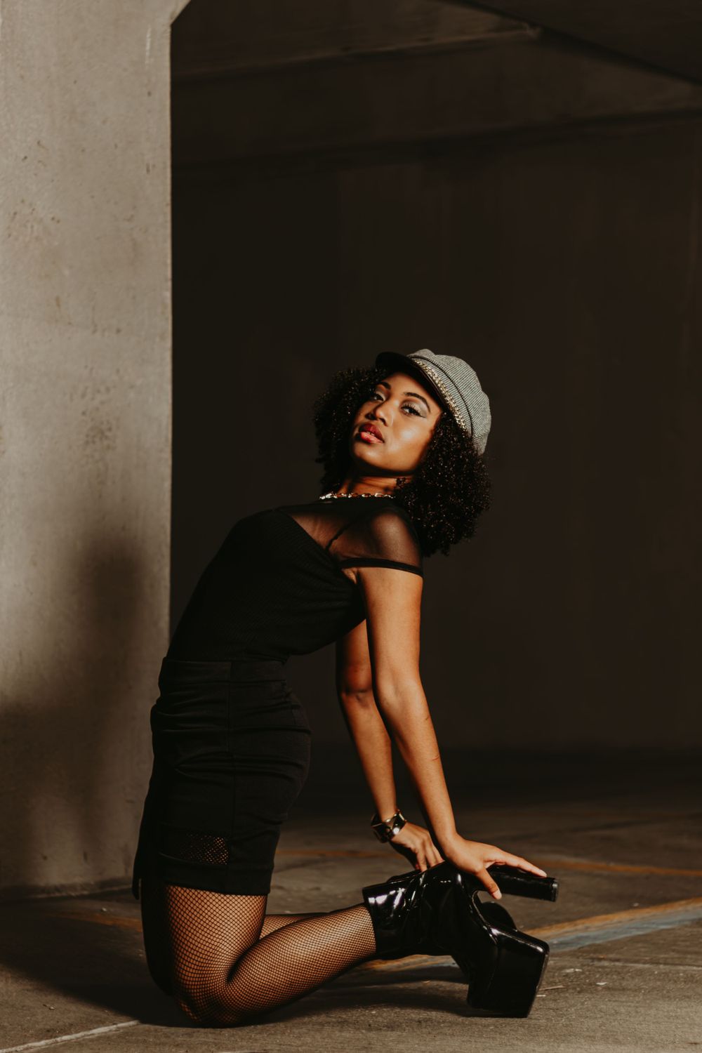 Model in black dress and cap poses elegantly on floor in dramatic lighting.