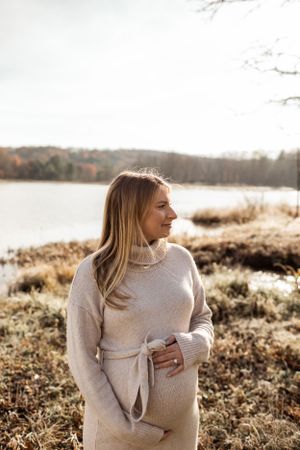 Person in cream sweater standing in autumn field near lake at sunset.