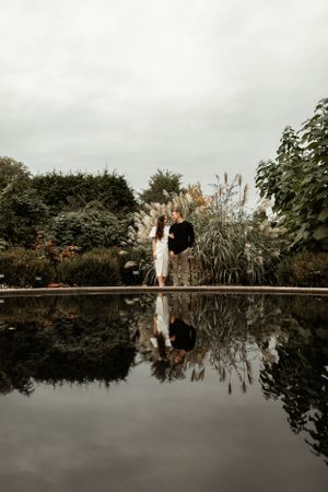 Two figures reflected in still water create a mirror image in a garden setting at dusk.