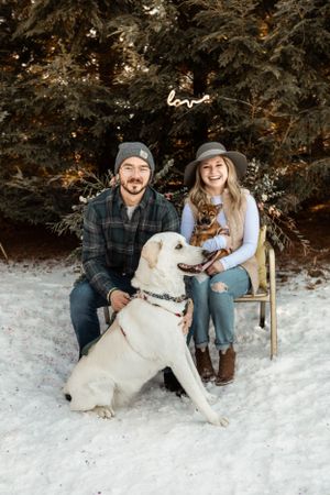 A couple and their large white dog pose together on a snowy winter day among evergreen trees.