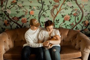 A couple sits on a brown couch against floral wallpaper in an intimate moment.