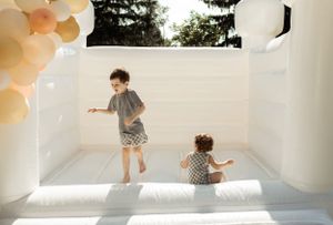 Two children jumping and playing in a white bounce house decorated with peach-colored balloons.