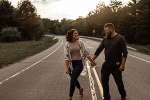 Couple walking and holding hands on empty road at sunset with trees in background.