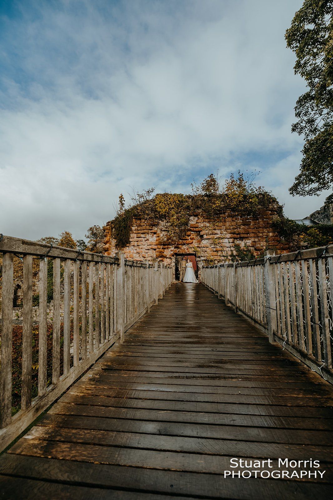 bride and groom stand under a stone archway at the end of the wooden bridge at Ruthin castle. Stuart Morris Photography.