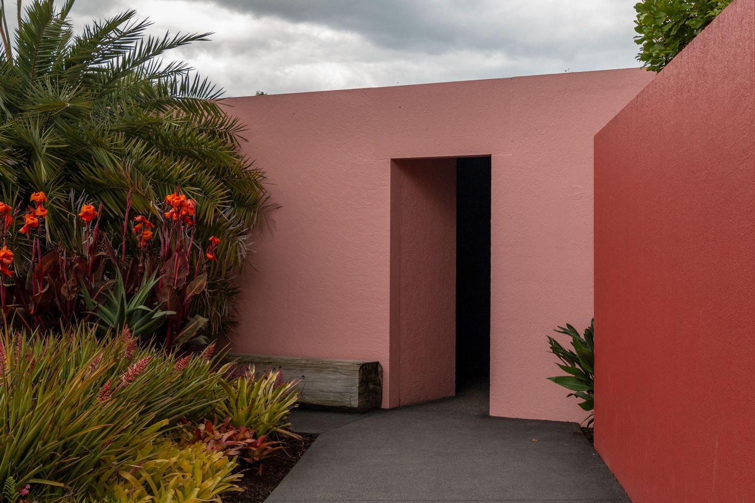 A modern courtyard entrance with smooth, pink-toned walls and clean architectural lines, surrounded by lush tropical plants and red flowers.