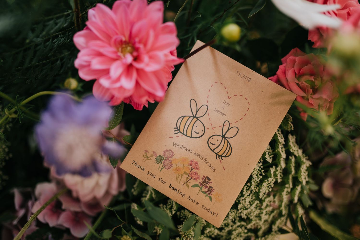 Cute bee illustration card nestled among pink and purple flowers in a garden setting.