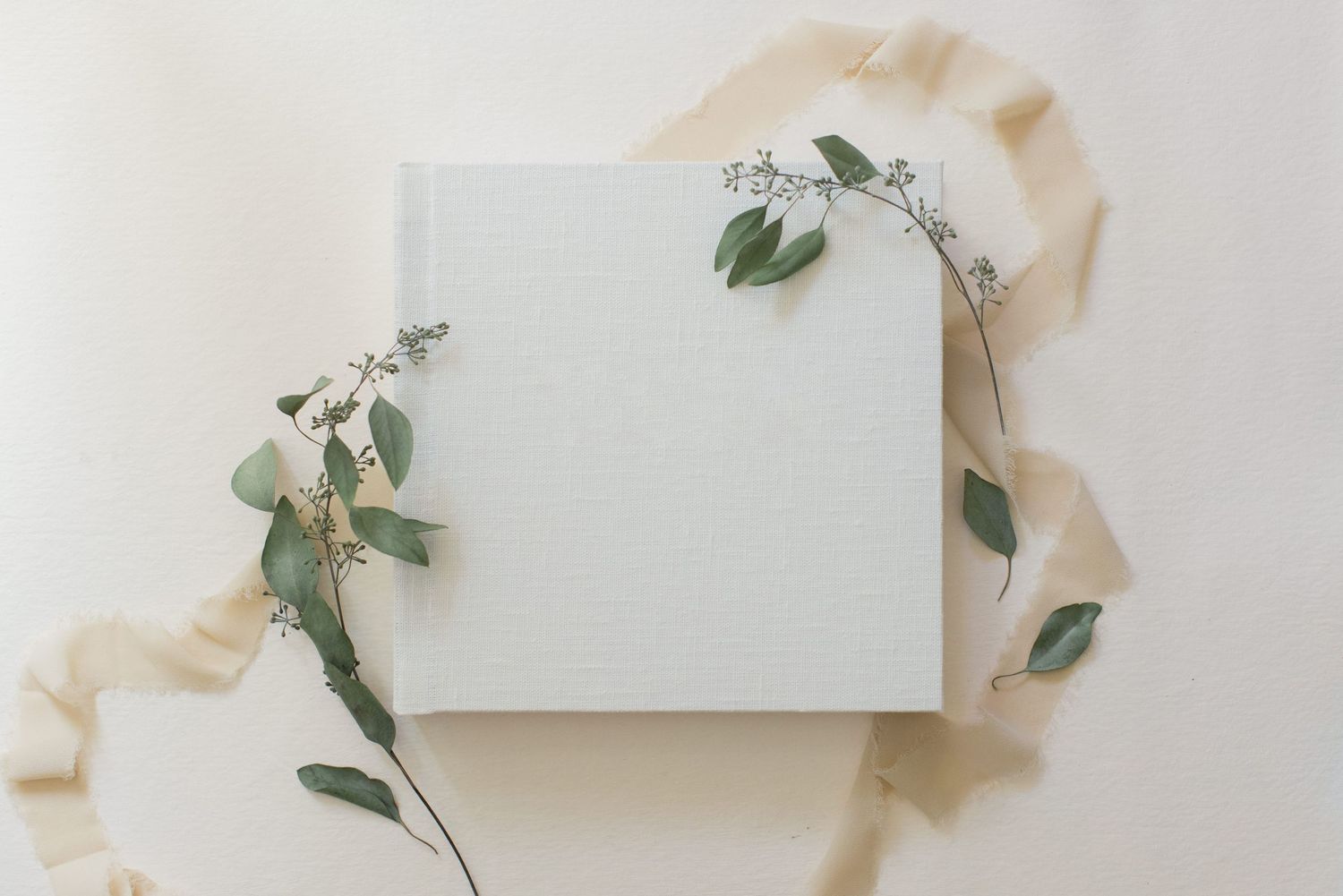 White photo album decorated with elegant eucalyptus sprigs on cream fabric background.