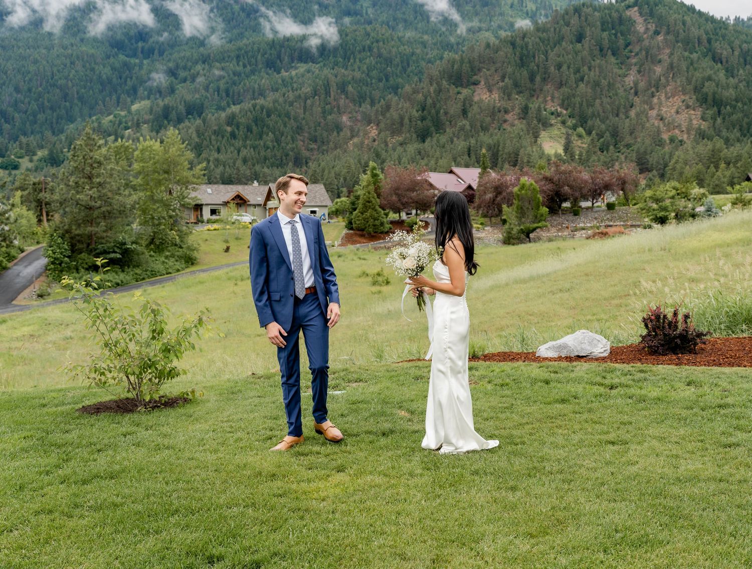 Couple in formal attire sharing a playful moment on grassy hillside with mountains in background.