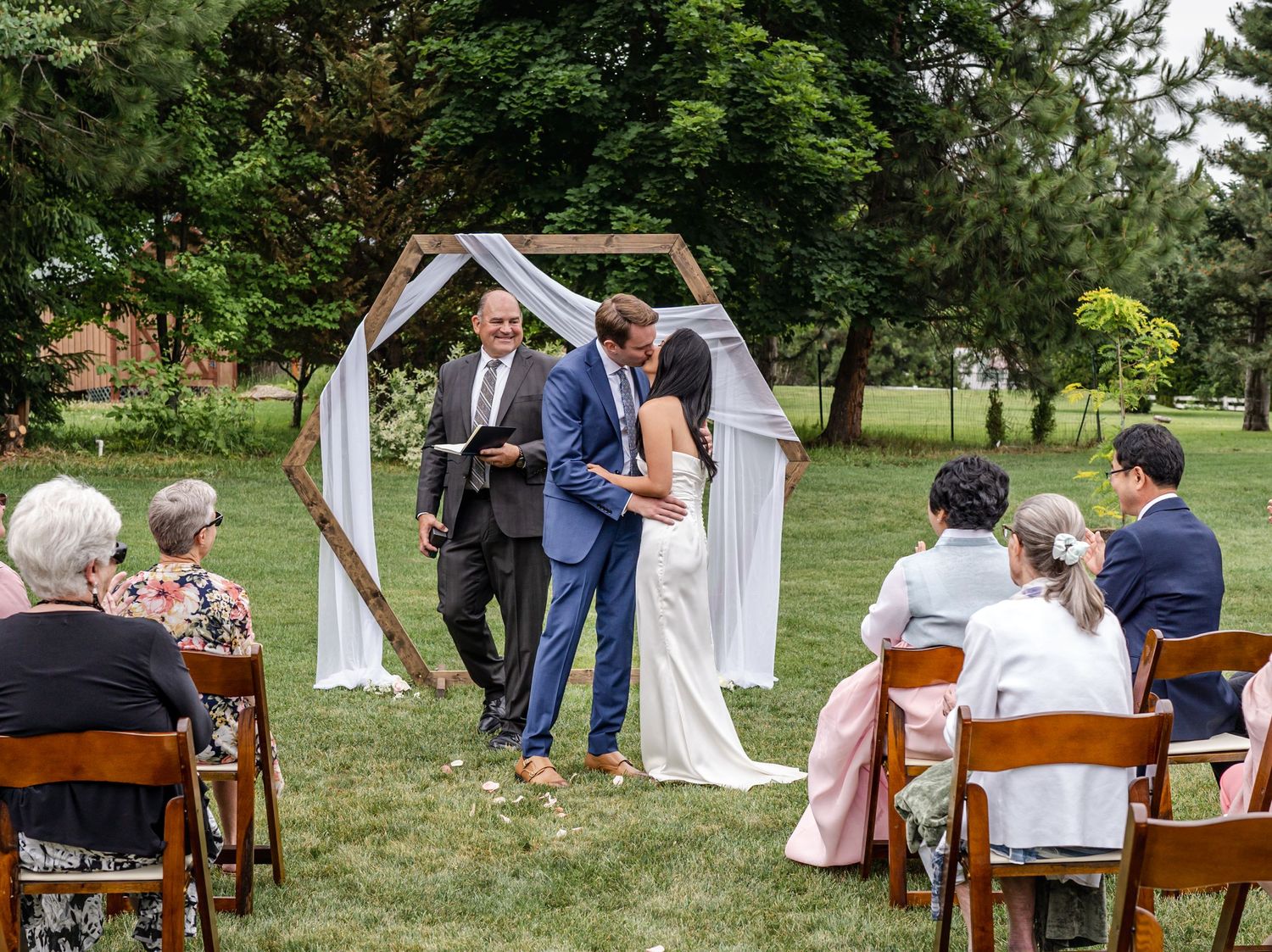 An outdoor wedding ceremony under a geometric arch with wooden chairs on a manicured lawn.