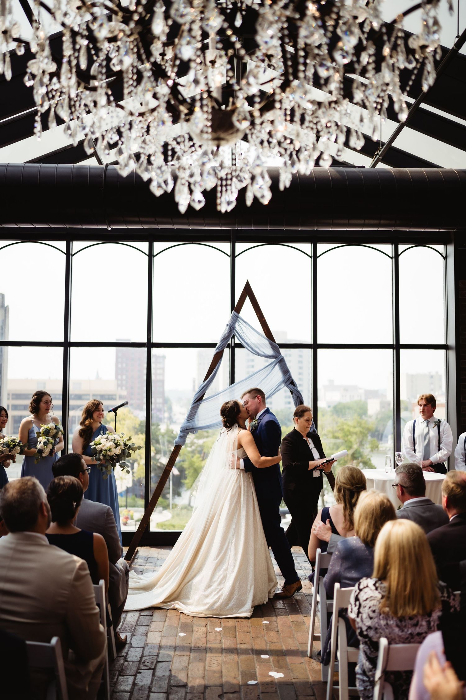 Wedding ceremony under crystal chandelier with geometric arch and floor-to-ceiling windows.