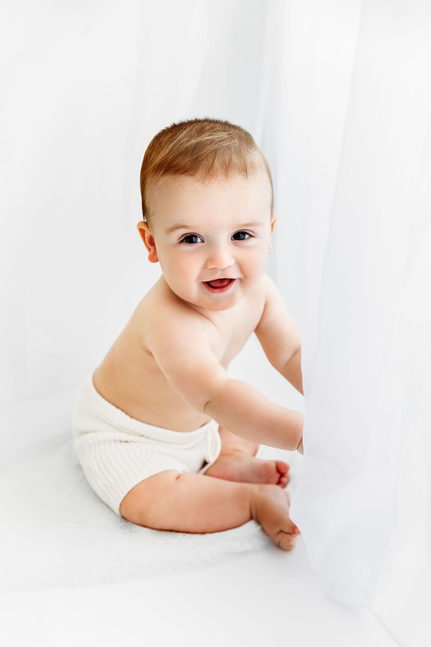 Happy baby playing with white curtains, wearing white knit shorts