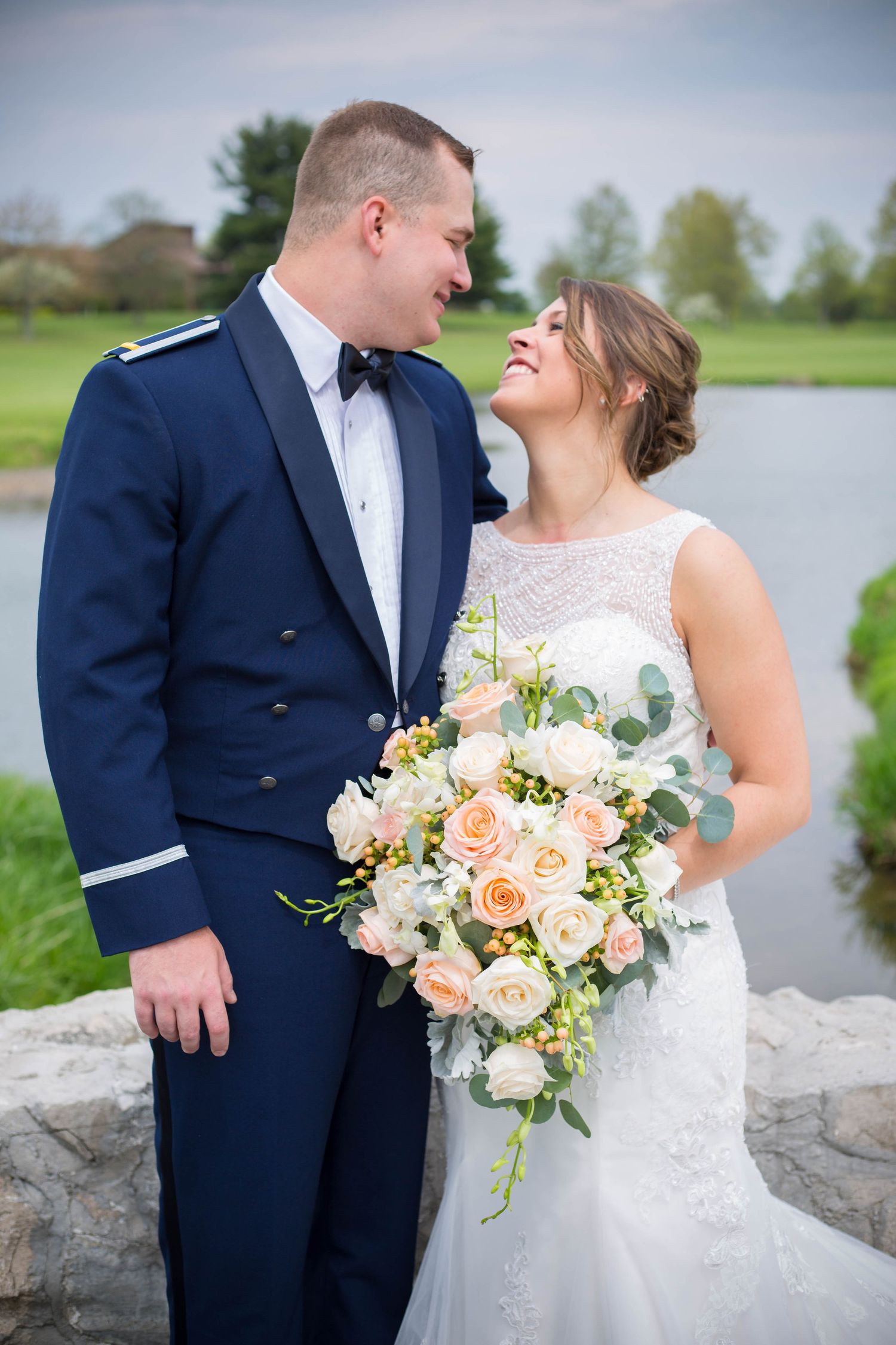 A couple shares a joyful moment by a lake, with the bride holding a peach and white floral bouquet.