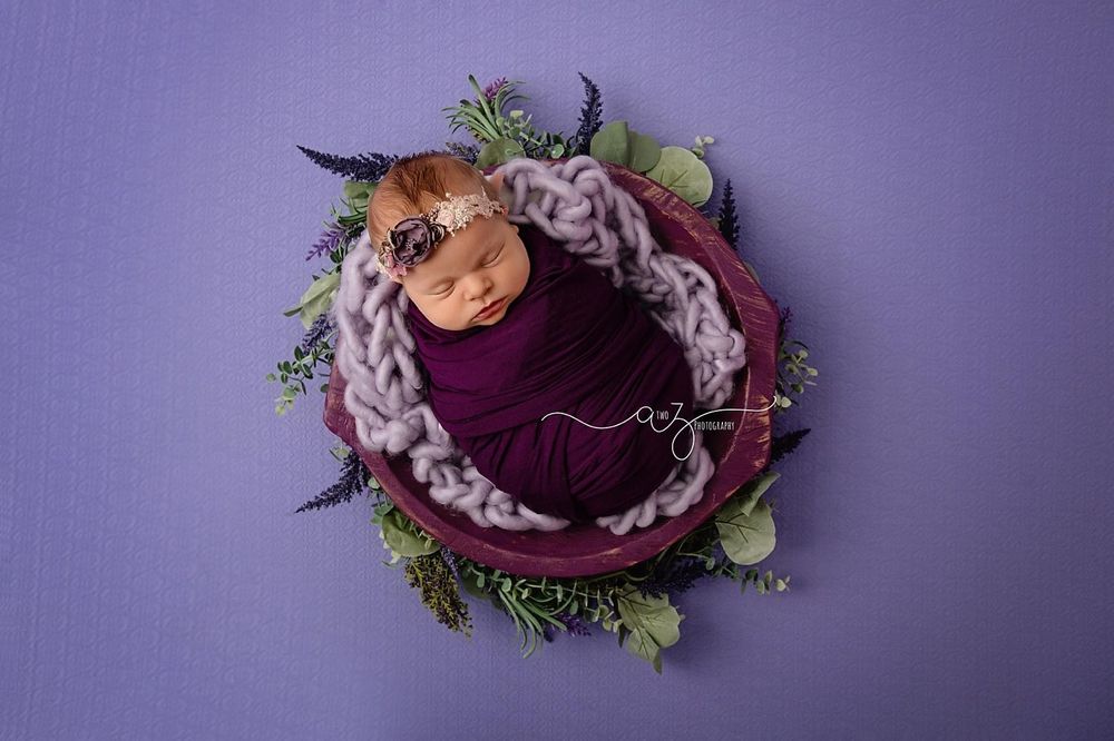 Newborn photography with baby nestled in purple fabric surrounded by floral elements.