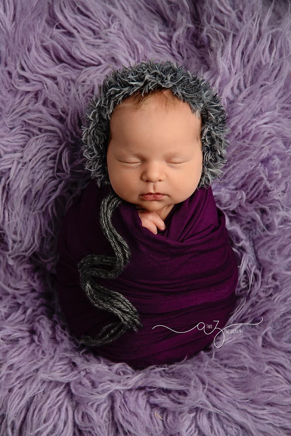 Sleeping newborn wrapped in purple fabric with gray bonnet on lavender fur backdrop.