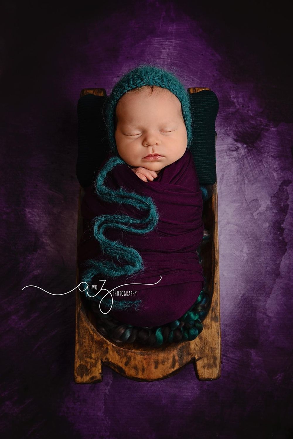 Artistic newborn portrait on wooden prop with teal accents against purple background.