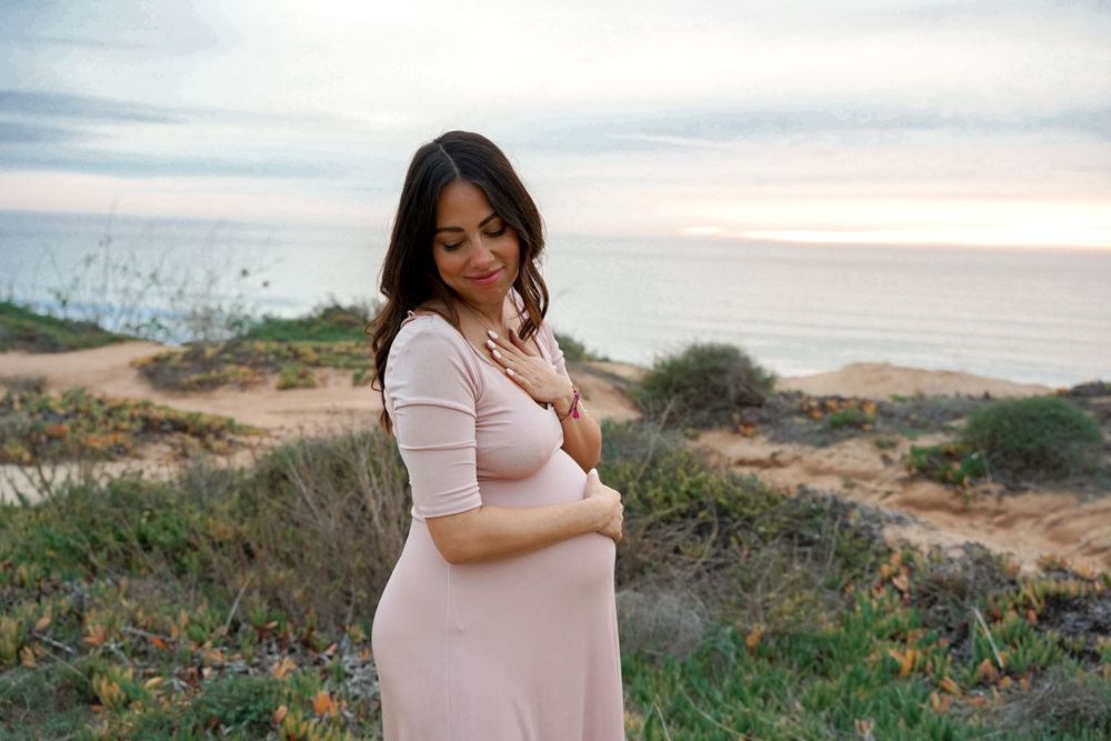 Expectant mother in pink dress stands thoughtfully on coastal cliffside during sunset.
