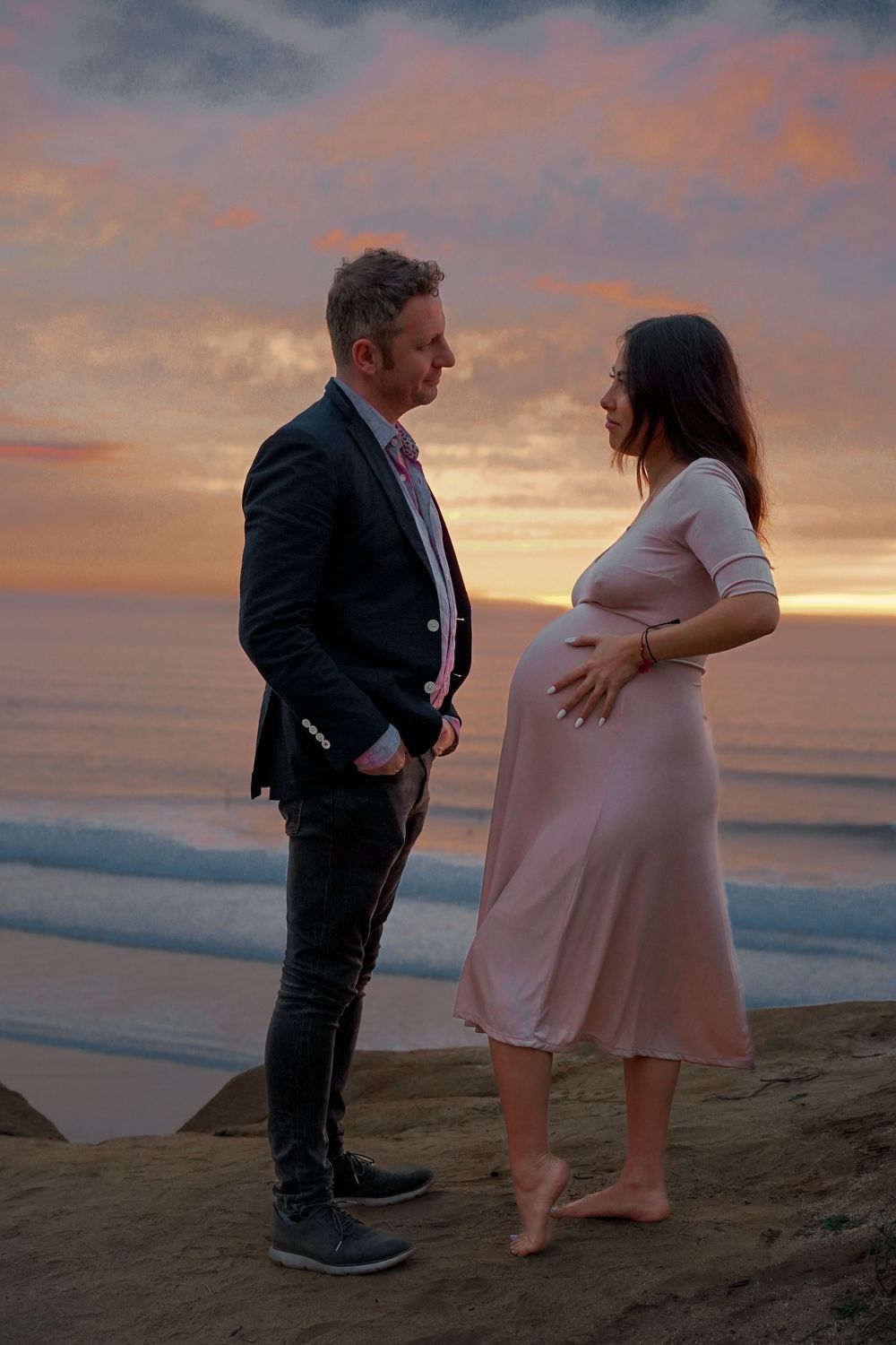 Sunset maternity photo with couple on beach cliff during golden hour.