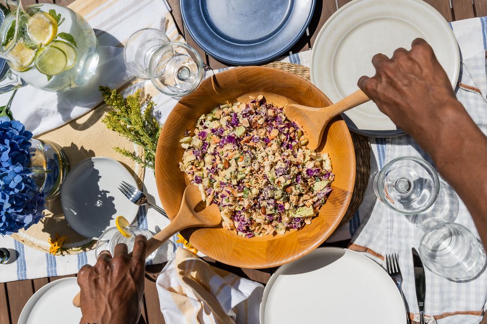 Food photography of an overhead view of hands reaching for a colorful quinoa salad bowl surrounded by table settings and wine glasses.