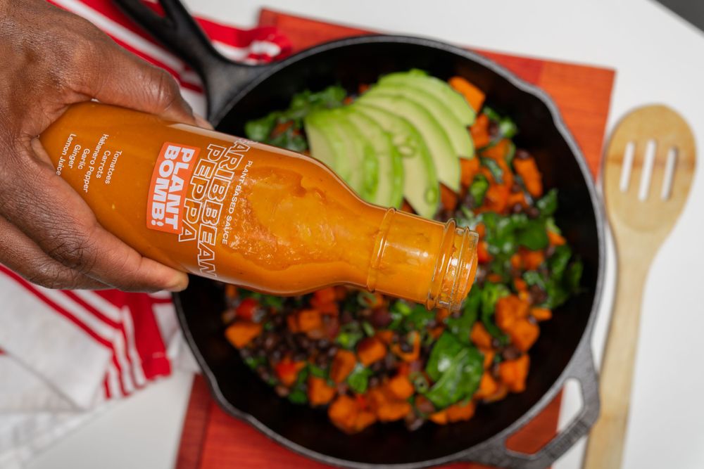 Food product photography of glass bottle being held over a black bowl filled with sweet potatoes, avocado, and fresh herbs.