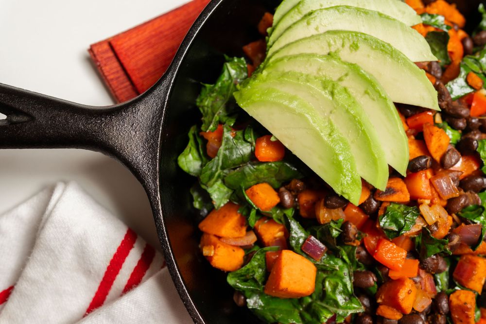 A cast iron skillet filled with roasted sweet potatoes, kale, and sliced avocado on a white cloth background. Food photography.