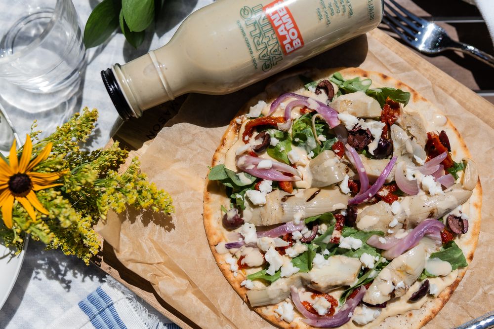 Food photography of a homemade pizza topped with artichokes, red onions and herbs sits on brown paper next to a wine bottle and sunflowers.
