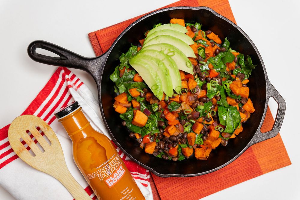 A cast iron skillet filled with sweet potato hash topped with fresh avocado slices and hot sauce bottle on the side. Food photography.