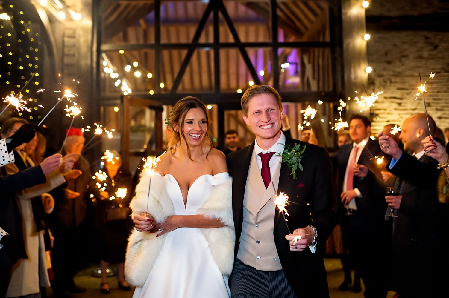 Couple celebrating during sparkler exit in rustic barn venue with festive lighting.