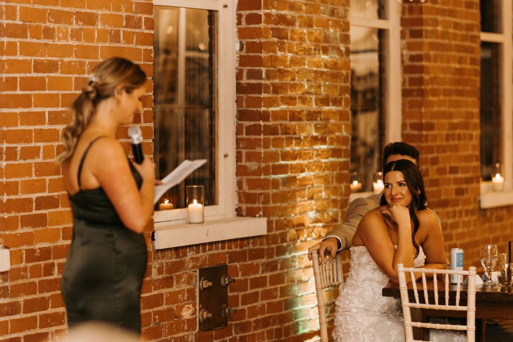 Bridesmaid reading heartfelt speech as emotional bride listens at wedding reception.