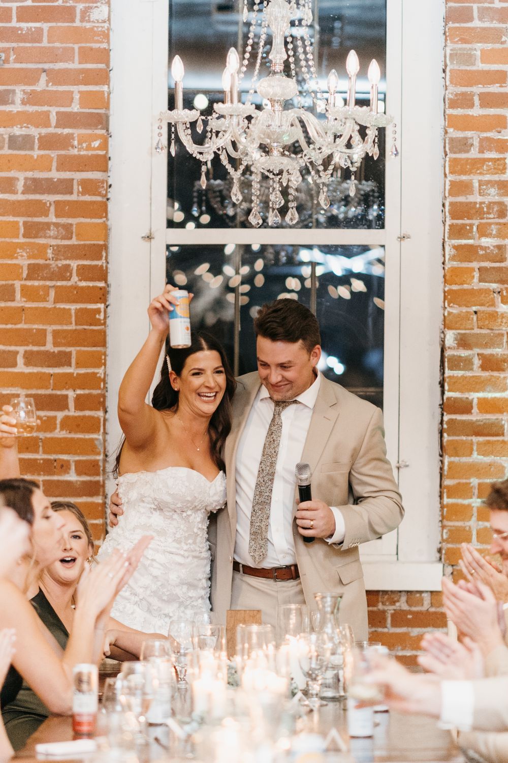 Laughing bride raises drink as groom gives speech at elegant wedding reception.