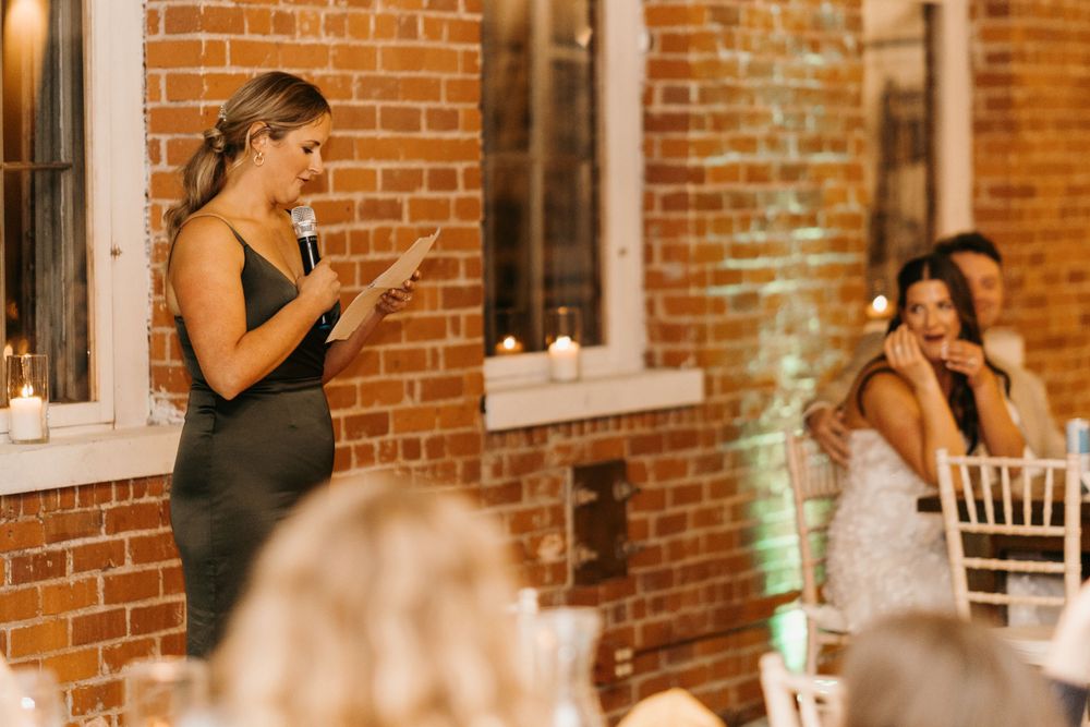 Woman in green dress giving wedding speech while bride smiles at brick venue.