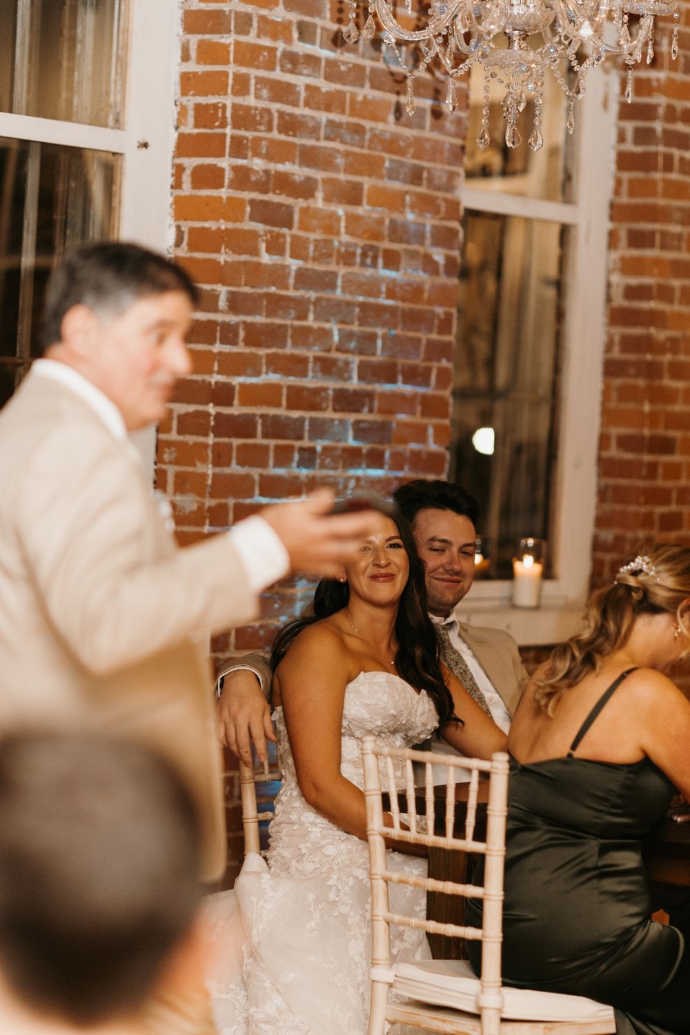 Bride laughing at wedding reception speech in rustic brick venue with chandelier.