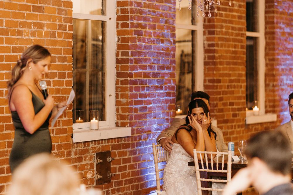 Bridesmaid delivering emotional wedding toast as bride wipes tears at reception.