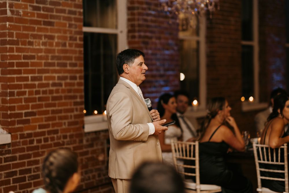 Elegantly dressed man in tan suit gives wedding speech to seated reception guests.