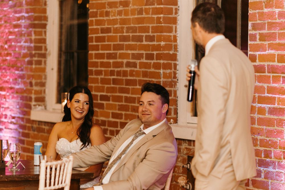 Bride and groom smiling during wedding reception speech at rustic brick venue.