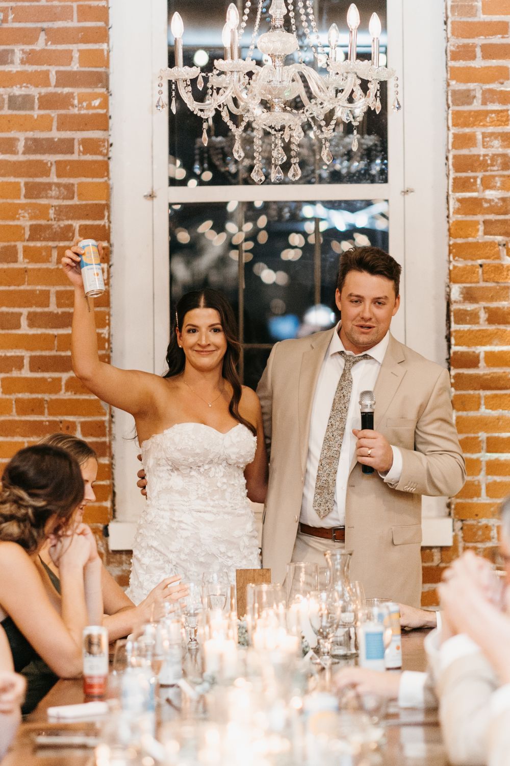 Bride raising can while groom speaks into microphone at wedding reception.