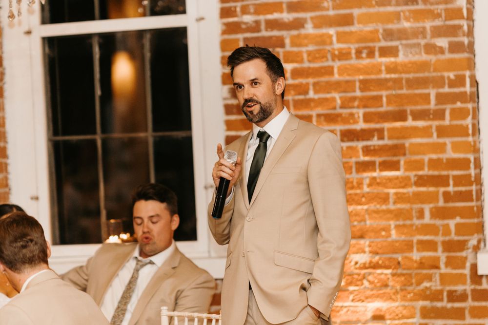 Man in tan suit giving wedding speech holding microphone at reception.