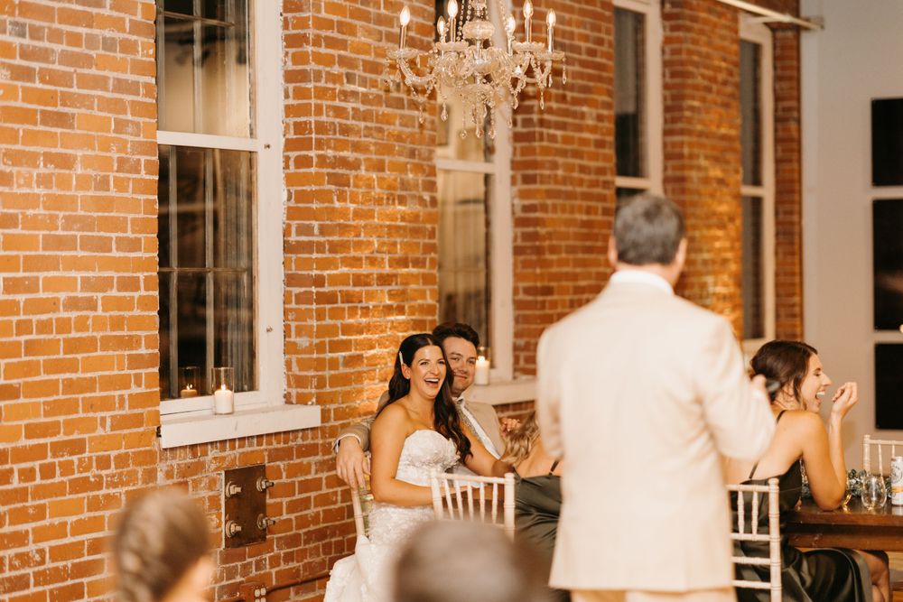 Bride and groom smiling during wedding reception toast in elegant brick-walled venue.