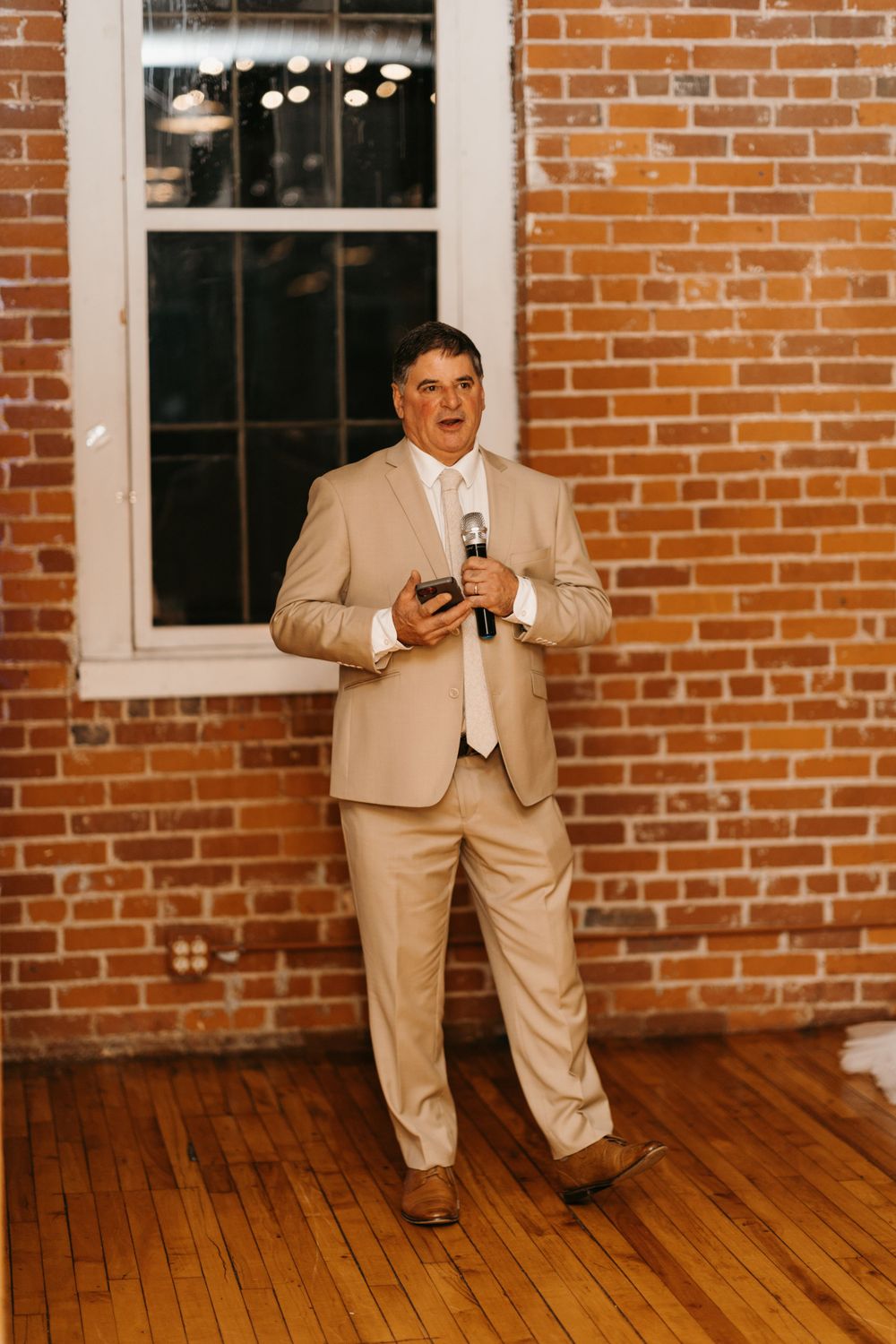 Man in tan suit delivers wedding speech holding microphone against exposed brick wall.