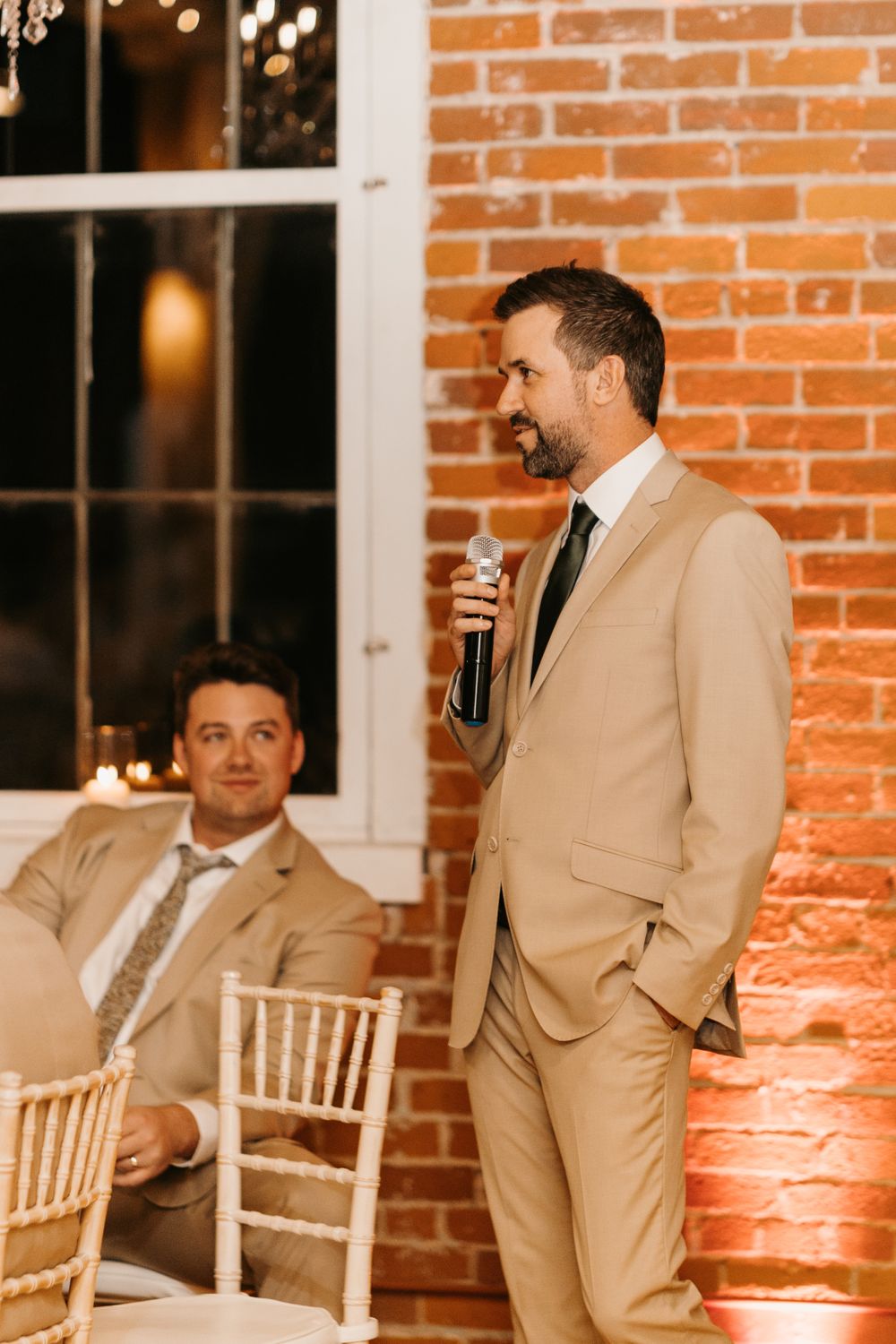 Groomsman delivering wedding toast with microphone in tan suit at brick venue.