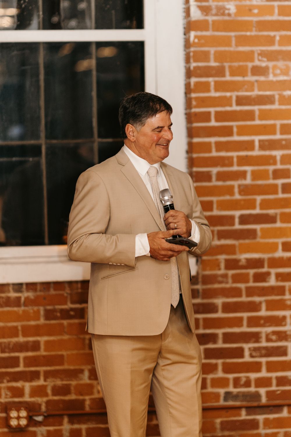 Smiling man in beige suit holds microphone while giving wedding toast by brick wall.