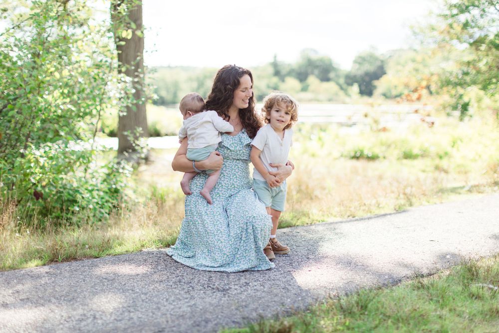 A person in a light blue maxi dress sits outdoors on a gravel path with two children in a sunny garden setting.