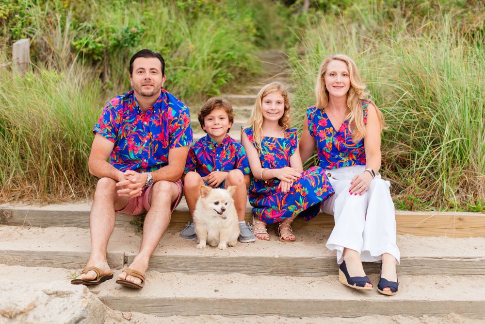 Family wearing matching blue floral outfits sits on concrete steps with their small white Pomeranian dog.
