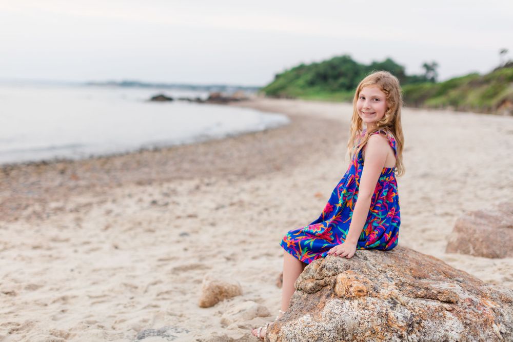 Woman in blue floral dress poses along sandy beach coastline at sunset with ocean waves in background.
