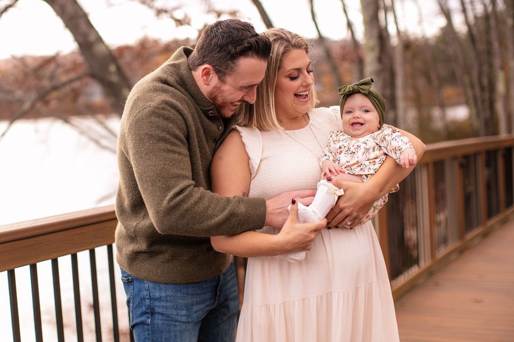 A couple shares a joyful moment with their baby while standing on a wooden bridge during sunset.