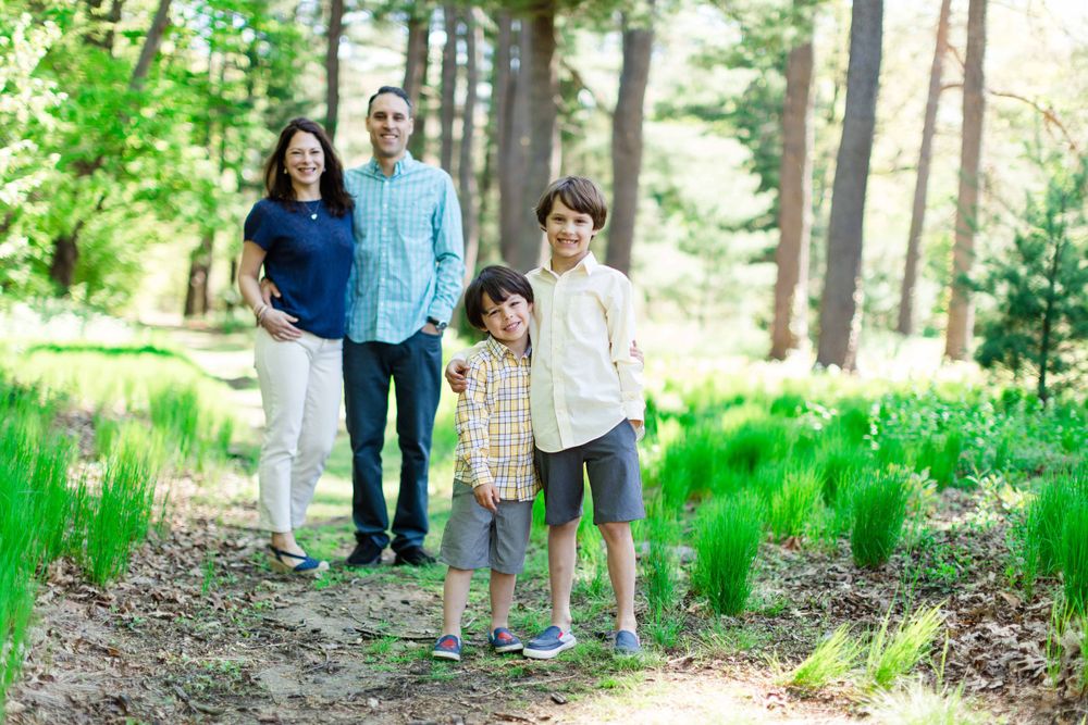 A family stands together on a wooded path surrounded by tall pine trees and green spring foliage.