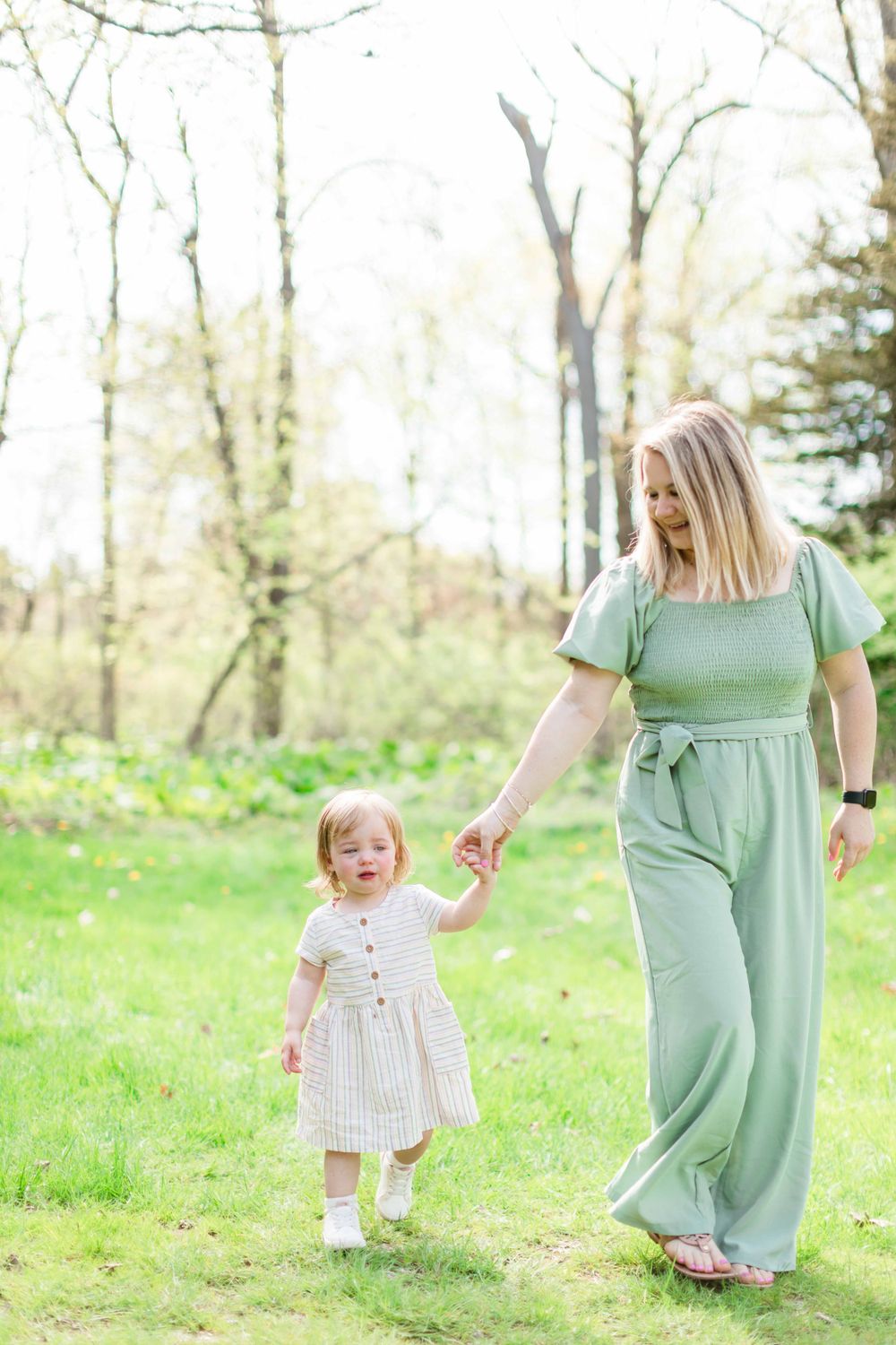 A little one wearing a white dress walking through a spring garden with tall trees in the background.