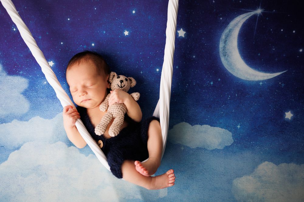 Sleeping newborn on swing prop against dreamy night sky backdrop with crescent moon.