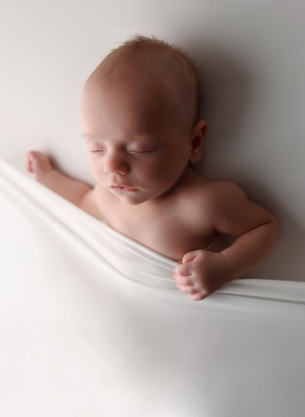 Artistic portrait of sleeping newborn wrapped in white fabric against light background.